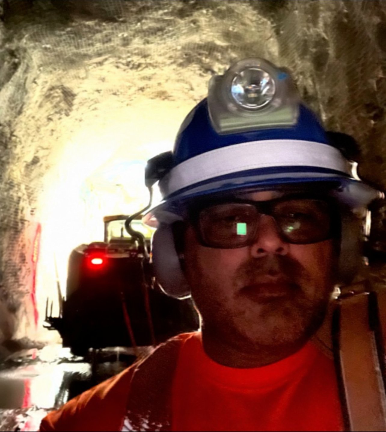 Jody Martinez underground in a blue hard hat with cap lamp, with a loader behind him